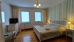 Een slaapkamer met een bed, een televisie, twee ramen, een kroonluchter en een nachtkastje met een lamp.