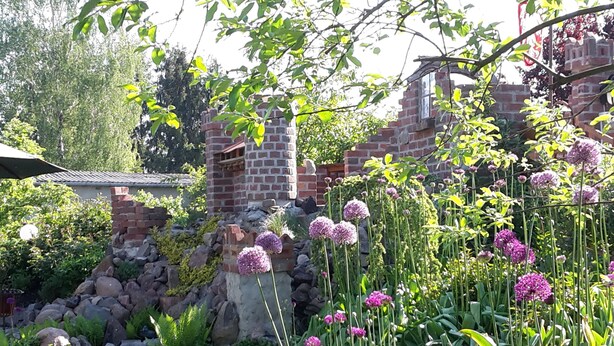 Een tuin met een bakstenen constructie, diverse planten en paarse bloemen.
