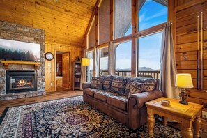 Ein gemütliches Blockhaus-Interieur mit Kamin aus Stein, einer Ledersofa, einem gemusterten Teppich und großen Fenstern mit Blick auf die Berge.