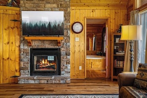 Ein gemütliches Wohnzimmer mit Kamin aus Stein, Holzwänden, einem Wand-Fernseher und einem Bücherregal.
