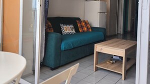 A living area with a teal sofa, a wooden coffee table, a white refrigerator, and a door leading to another room. - Grand studio lumineux pour 2 ou 4 personnes (Ducos)