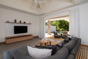 Uma sala de estar moderna com televisão de tela plana, um sofá cinza, uma mesa de centro de madeira e vista para área externa com piscina e vegetação.