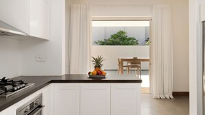 Una cocina moderna con armarios blancos, encimera negra y estufa a gas. Hay un frutero en la encimera y vistas a un balcón con plantas.