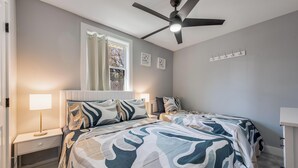 Quarto com duas camas, um ventilador de teto, janela com cortinas, abajures e um cabideiro fixado na parede.