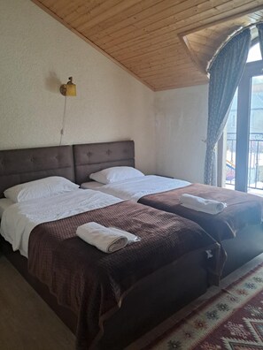 A room with two beds, a wooden ceiling, a window with curtains, and a wall-mounted lamp. - OTEL IVERIA (Stepantsminda)