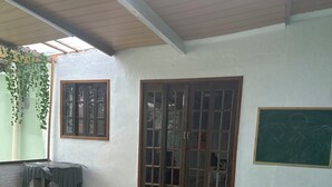 A covered patio area with a tiled floor, a green artificial turf section, a wooden door, a window, and a chalkboard on the wall. - Pousada simples  (Pinhais)