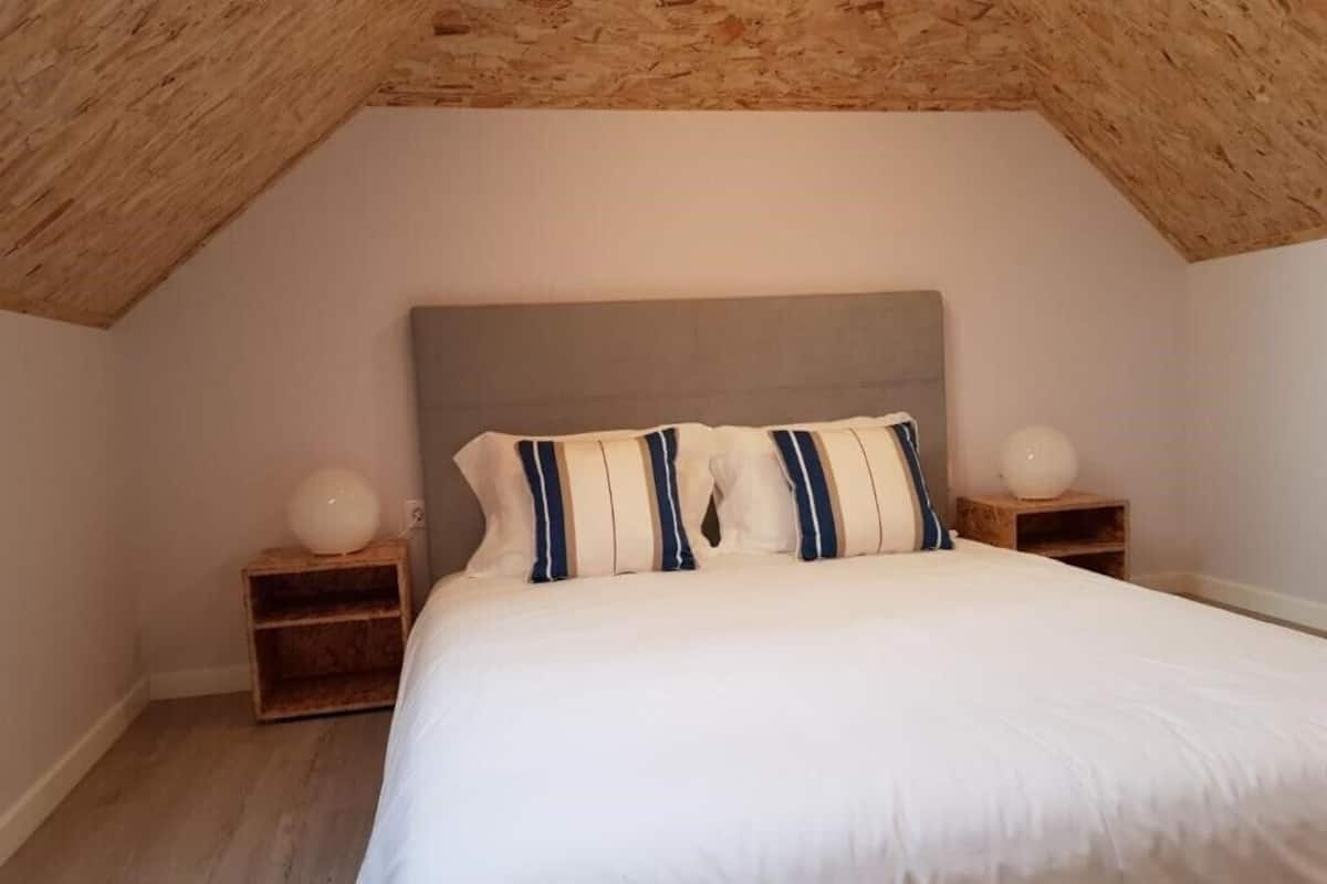 A bedroom with a sloped ceiling, a bed with white bedding and striped pillows, two bedside tables with lamps, and a wooden headboard.
