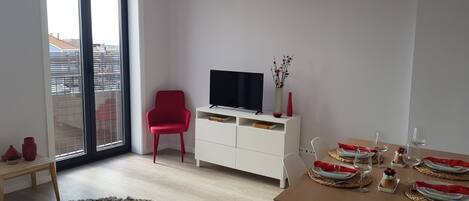 A modern living room with a large window, a red chair, a white TV stand, and a dining area set with plates and glasses.