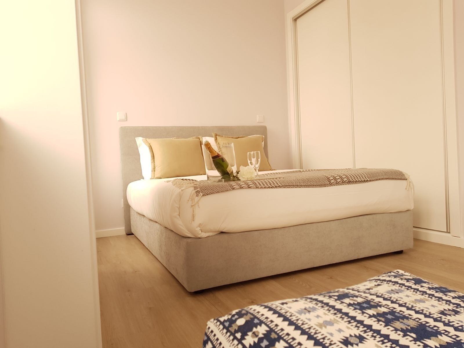 A bedroom with a bed, pillows, a bedside table with a bottle and glasses, a patterned rug, and a wardrobe.