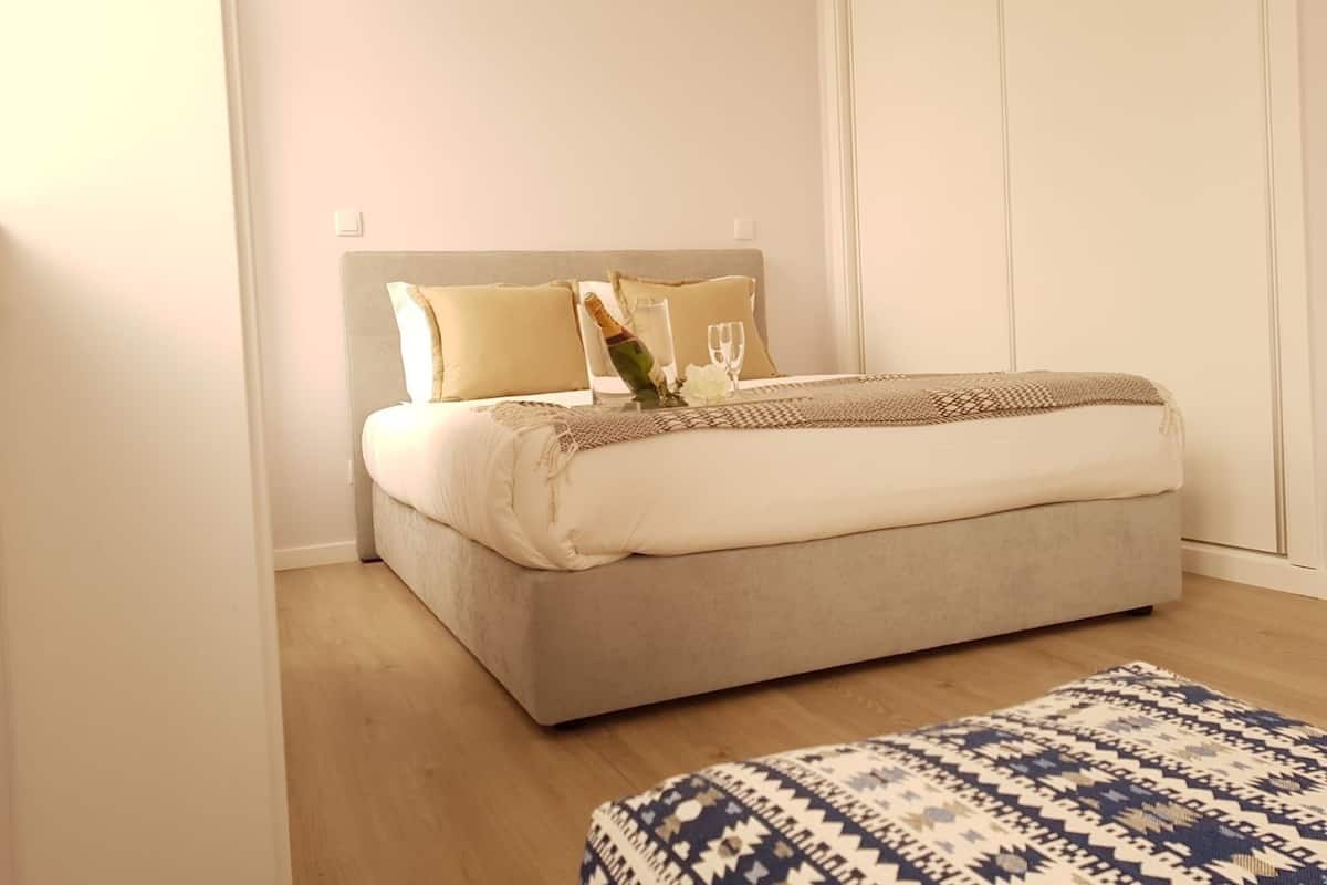 A neatly made bed with a grey headboard, a bedside table with a bottle and glasses, and a patterned rug on the floor.