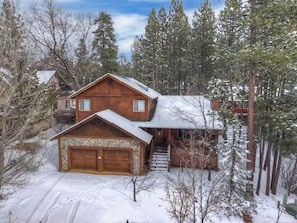 A two-story wooden cabin with a stone foundation, a garage, and a staircase leading to a porch. - Alpine Woods Log Cabin•4BR 3BA•Spa&Mountain Views (Big Bear Lake)