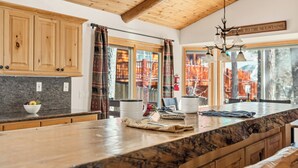Une cuisine avec des meubles en bois, un plan de travail en marbre et une vue sur un chalet de montagne, visible à travers de grandes fenêtres.