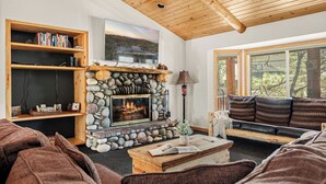 Un salon chaleureux avec une cheminée en pierre, des étagères en bois, une grande fenêtre et un coin salon confortable.