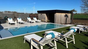 A pool area with lounge chairs, a small building, and wind turbines in the background. - Inspire Residence and Wellness (Tongeren-Borgloon)