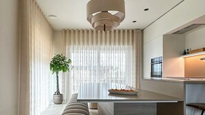A modern kitchen with a central island, a dining area with a bench, and a large window with curtains. - Inspire Residence and Wellness (Tongeren-Borgloon)