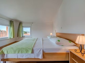 Une chambre d’hôtel avec deux lits, une fenêtre avec des rideaux, une table de chevet avec une lampe, et une vue sur les bâtiments environnants.