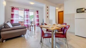 A modern living room with a sofa, dining table, and chairs, a kitchen area with a refrigerator, and a balcony with curtains. - Mise Holiday Apartments - Two bedroom apartment II (Kastel Gomilica)