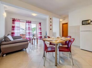 A modern living room with a sofa, dining table, and chairs, a kitchen area with a refrigerator, and a balcony with curtains. - Mise Holiday Apartments - Two bedroom apartment II (Kastel Gomilica)