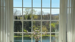 A room with a large window, a desk with a laptop, a chair, and a vase with flowers on the window sill. - Hudson Valley Modern 5 BR Farmhouse with Pond and Stunning Sunsets (Pine Plains)