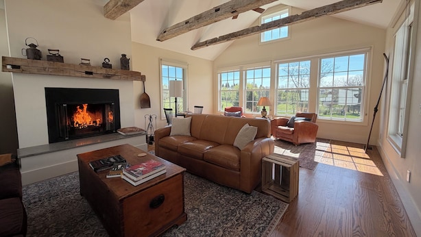 A spacious living room with a fireplace, wooden beams, large windows, and a cozy seating area. - Hudson Valley Modern 5 BR Farmhouse with Pond and Stunning Sunsets (Pine Plains)