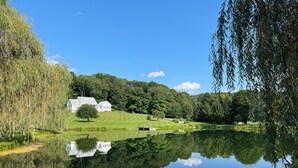 A serene landscape with a pond reflecting a white building and trees. - Hudson Valley Modern 5 BR Farmhouse with Pond and Stunning Sunsets (Pine Plains)