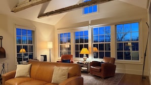 A living room with a leather sofa, two armchairs, a coffee table, and a bookshelf. There are large windows with multiple panes, a wooden ceiling beam, and a lamp on the coffee table. - Hudson Valley Modern 5 BR Farmhouse with Pond and Stunning Sunsets (Pine Plains)