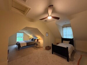 Ein Schlafzimmer mit schrägem Dach, Deckenventilator, zwei Betten, einem Fenster mit Vorhängen und einem kleinen Nachttisch mit Lampe.