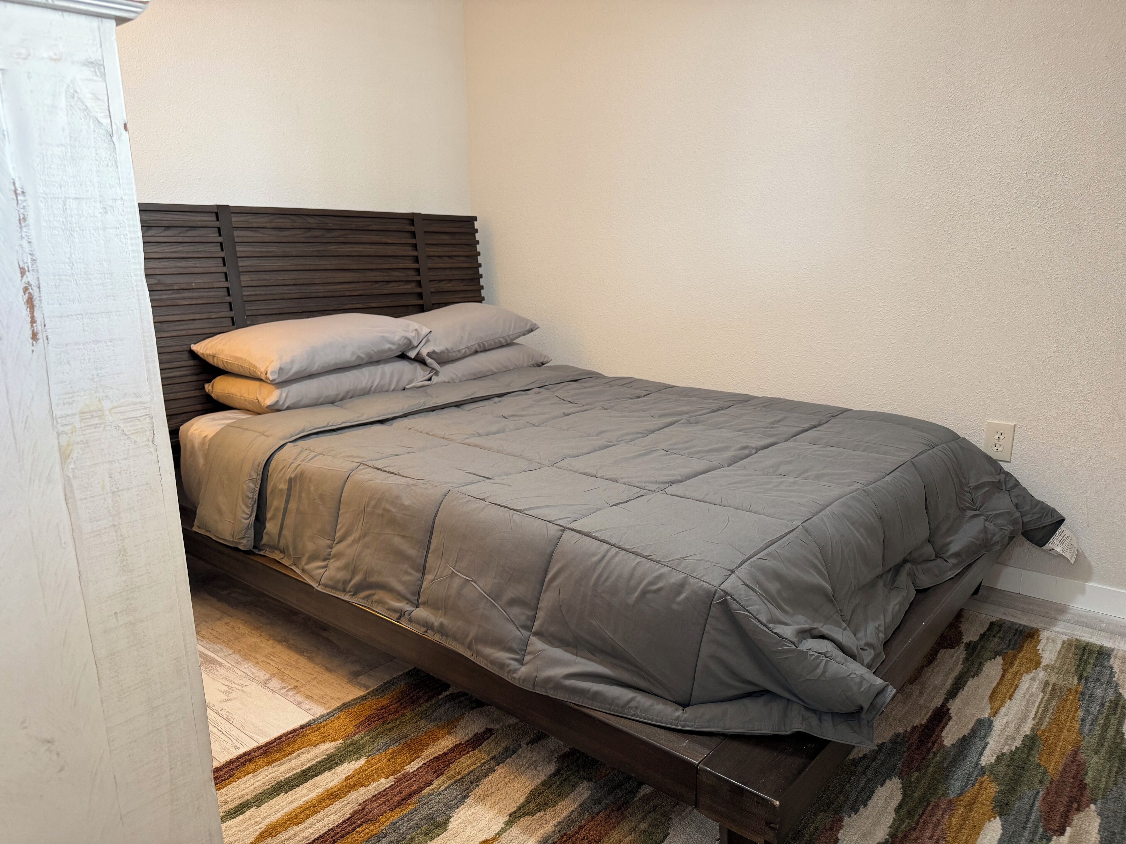 A single bed with a wooden headboard, a striped rug, and a plain wall.