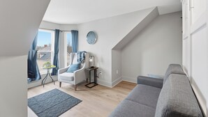 Een moderne woonkamer met een grijze bank, een klein bijzettafeltje, een stoel, een klok en uitzicht op het buitenlands landschap door een raam met blauwe gordijnen.
