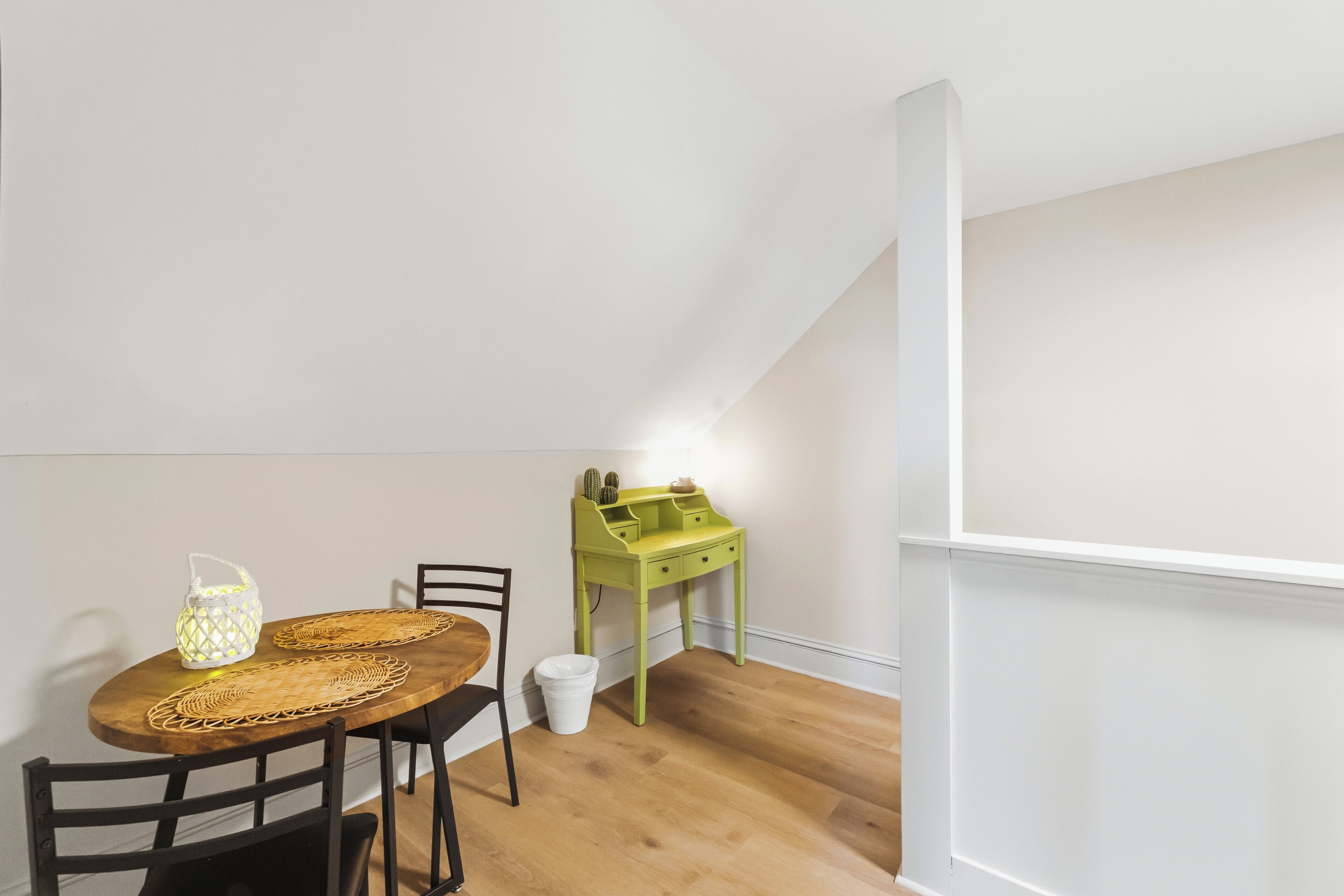 A room with a wooden dining table, chairs, a small green desk, a white trash bin, and a white wall.
