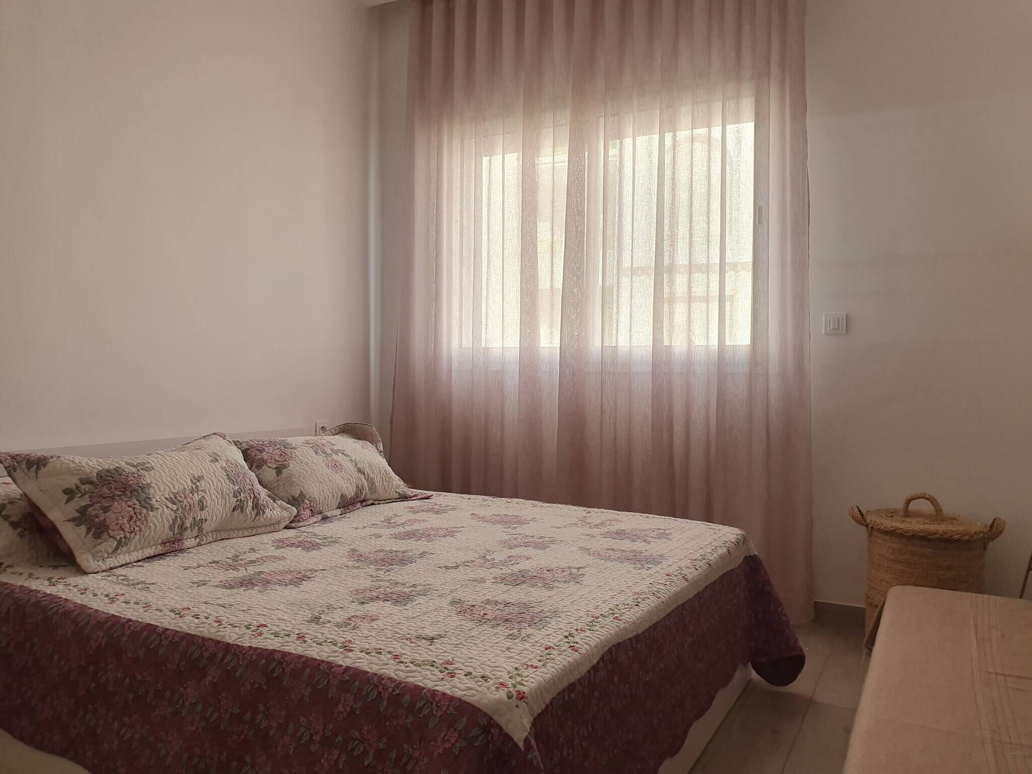 A bedroom with a bed, floral bedding, a window with sheer curtains, and a small wicker basket.