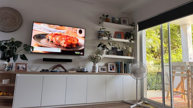Un soggiorno con televisore, mobili bianchi, un ventilatore e un'ampia finestra vista sul verde.