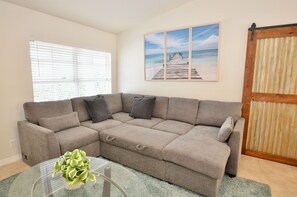 A beige sectional sofa, a glass coffee table with a plant, a wooden door, and a beach scene painting on the wall.