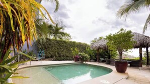 Une piscine à bord incurvé, entourée de plantes tropicales et bordée d’une structure à toit de chaume.