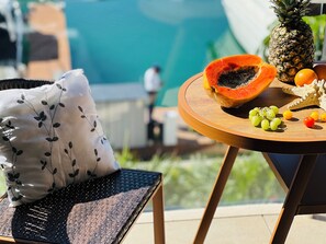 A poolside setting with a wicker chair, a round wooden table with fruits, and a white pillow with a leaf pattern. - MARINA CONDO CORAZON (Cancún)
