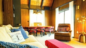 Une pièce chaleureuse avec un plafond en bois, un coin repas avec des chaises rouges, un fauteuil en cuir marron et une table recouverte d’une nappe rouge.