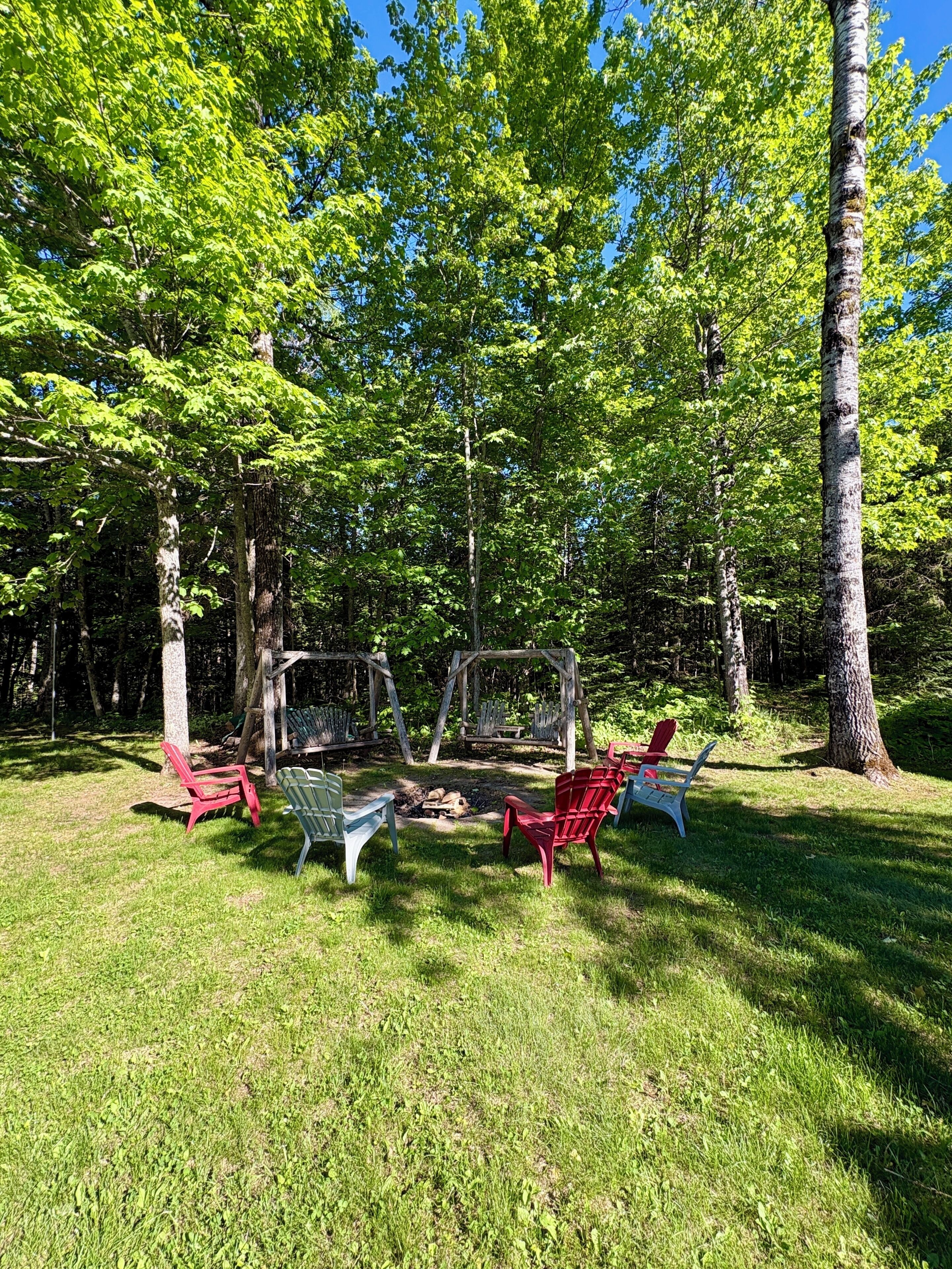 A forested area with a fire pit and Adirondack chairs.
