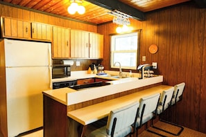 A kitchen with wooden cabinets, a refrigerator, a microwave, a sink, and a bar area with chairs.