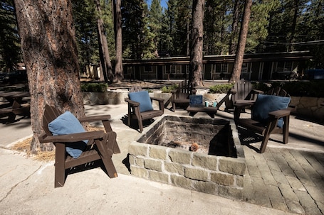 A fire pit surrounded by wooden chairs and benches, set in a wooded area with trees and a building in the background.
