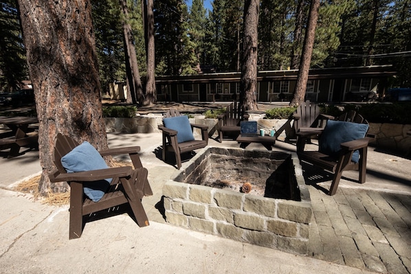 A fire pit surrounded by wooden chairs and benches, set in a wooded area with trees and a building in the background.