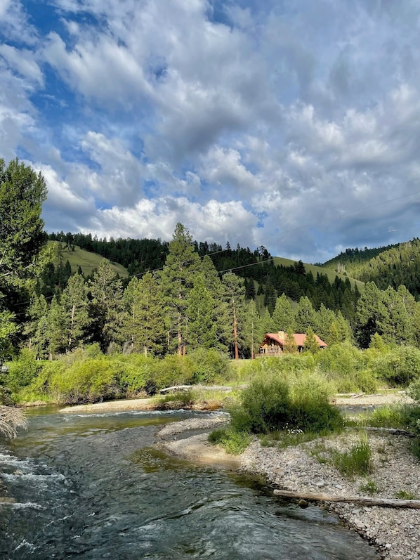 Vista a la orilla del río con una cabaña rodeada de bosque denso y colinas.