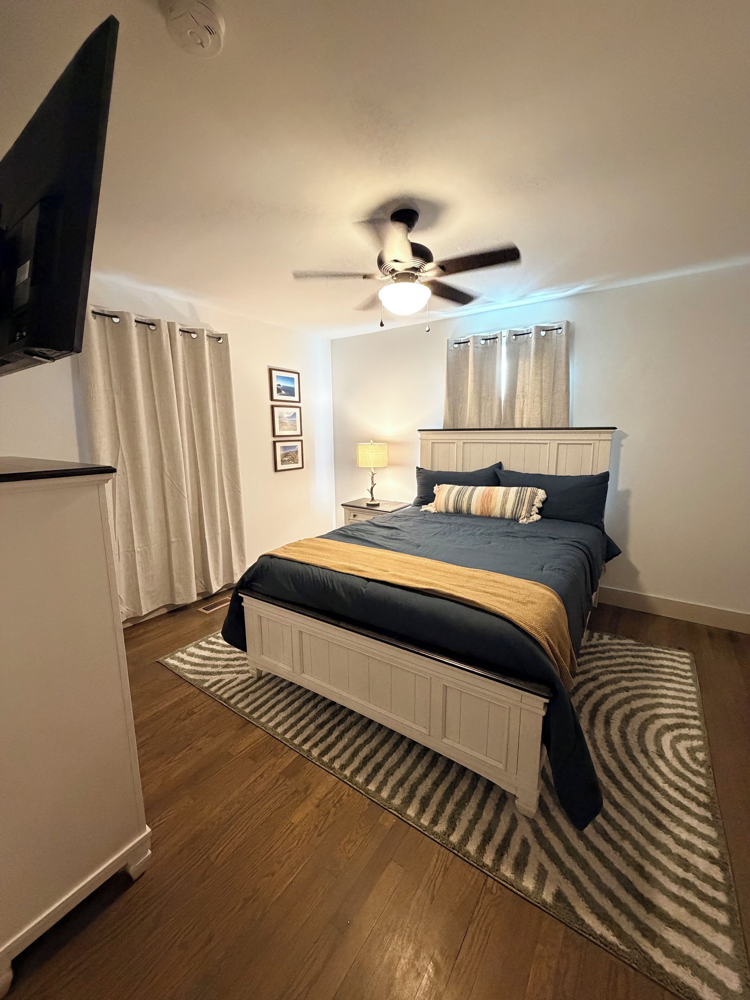Une chambre à coucher avec un lit, un ventilateur de plafond, une télévision et une table de chevet avec une lampe.