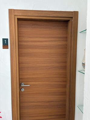 A wooden door with a metal handle and a keyhole, a number sign on the wall, and a glass shelf with a white object. - Noonbir Alaçatı (Cesme)