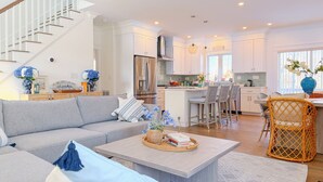 Una cucina moderna con isola centrale, zona pranzo con sedie in vimini, un divano grigio con cuscini decorativi e una scala che conduce a un altro livello.