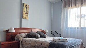 Siististi pedattu sänky, jossa on kuviollinen päiväpeite, kaksi yöpöytävalaisinta, puinen yöpöytä ja taulu seinällä.