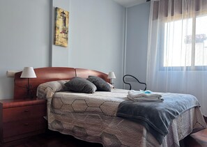 A neatly made bed with a patterned bedspread, two bedside lamps, a wooden nightstand, and a painting on the wall. - Golfiño Apartment (O Grove)
