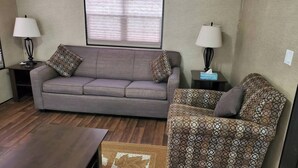 Un salón con un sofá gris, un sillón estampado, dos mesitas de noche con lámparas y una ventana con persianas.