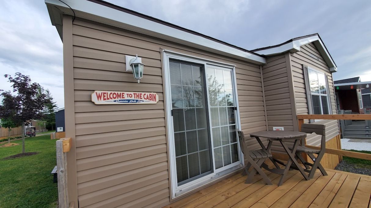 A cabin with a 'Welcome to the Cabin' sign, a sliding glass door, and a small patio with a table and chairs.
