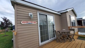 Una cabaña con un letrero que dice 'Welcome to the Cabin', una puerta corrediza de vidrio y un pequeño patio con mesa y sillas.