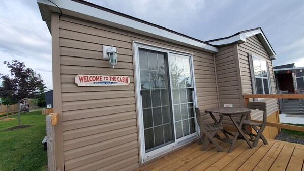 A cabin with a 'Welcome to the Cabin' sign, a sliding glass door, and a small patio with a table and chairs.